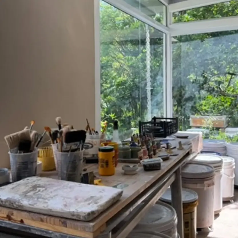 Mesa de trabalho do ateliê com pincéis e esmaltes, ao lado de janelas para o jardim
